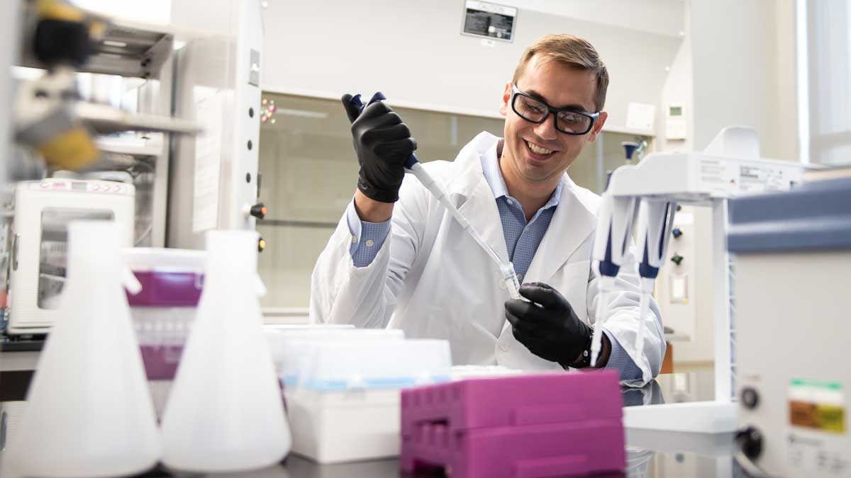 A man in a lab coat, gloves and safety glasses pipes liquid into a small plastic container.