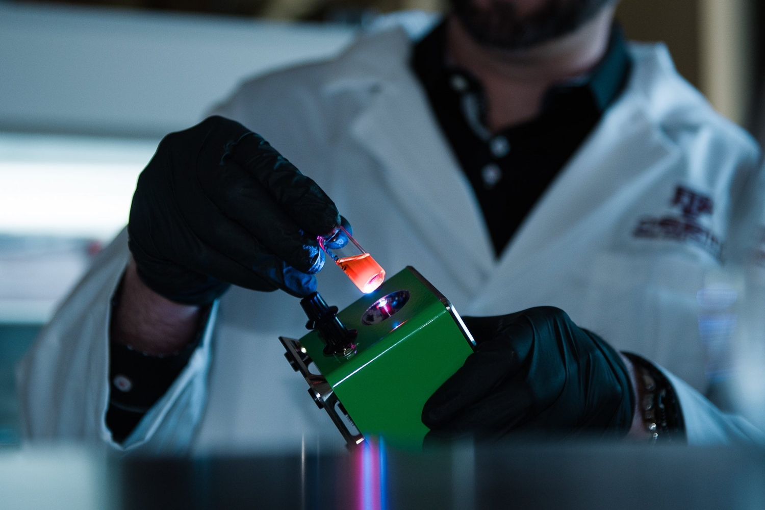 Close-up of a chemist wearing a while lab coat and black gloves while holding a glass vial containing a fluorescent material that is glowing bright orange due to its exposure to an ultraviolet light source within the lab