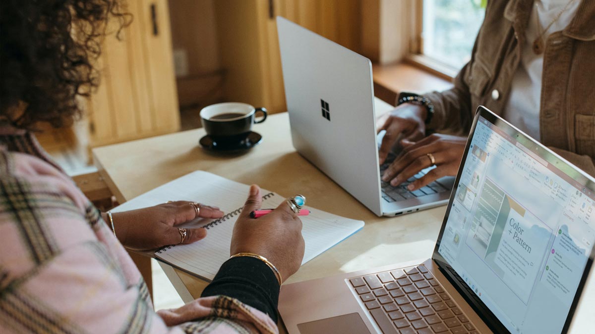 Two people sit at a table, writing in a journal and working on their laptops