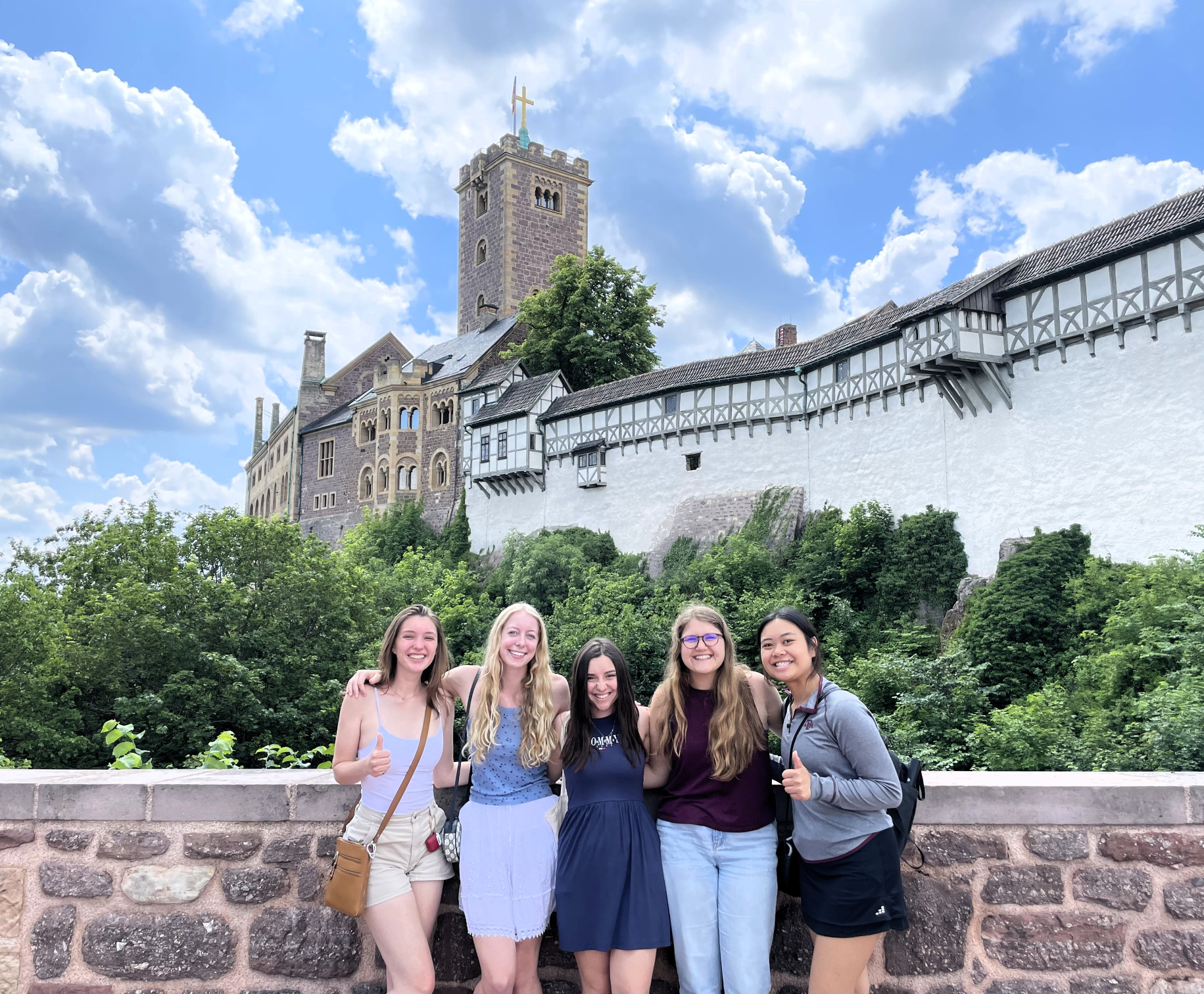 Five people stand on a stone bridge in front of a historic hilltop castle featuring a tall central tower with a cross, white walls, timber framing, and stone architecture. The surrounding landscape is lush and green under a bright, partly cloudy sky, suggesting a pleasant day. 