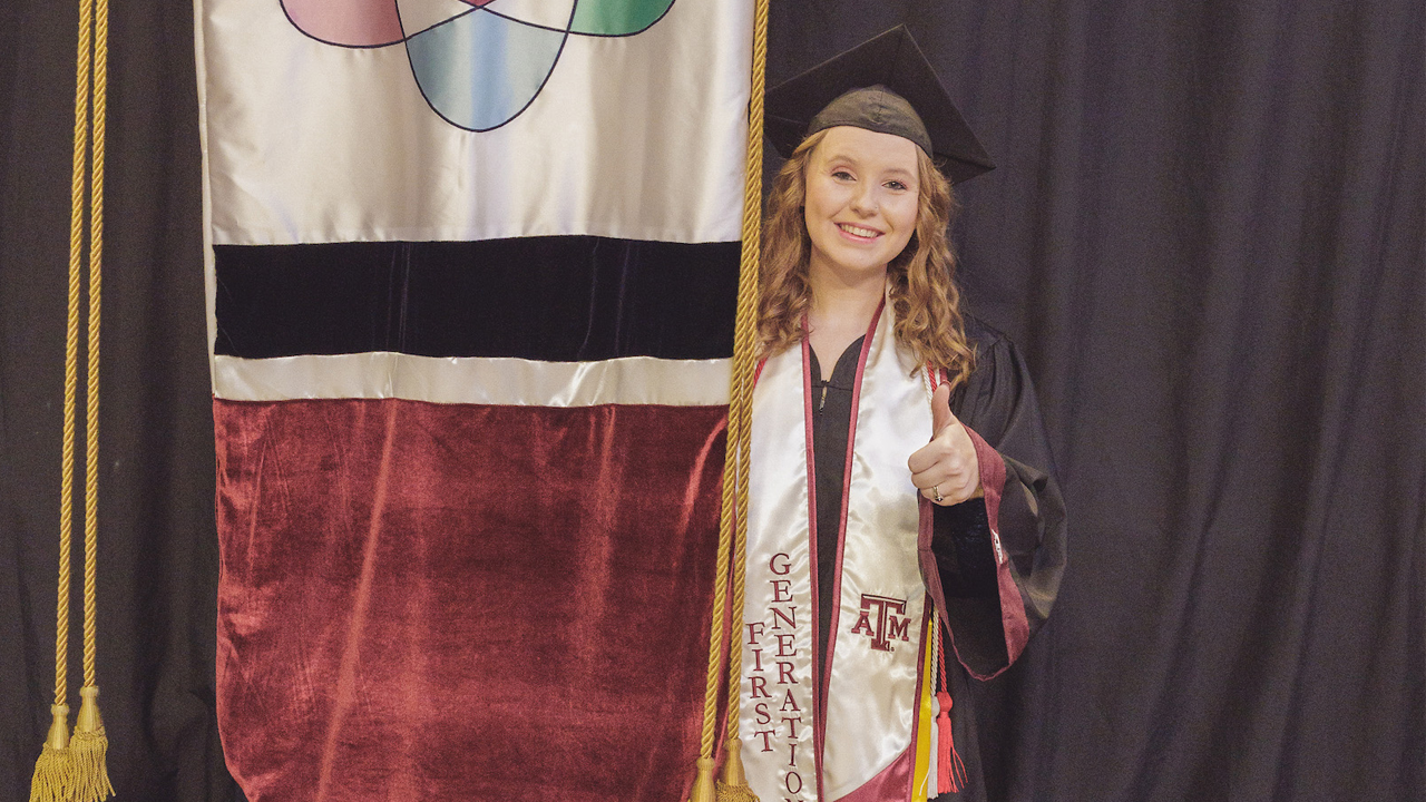 Megan in a black graduation gown and cap smiling proudly, wearing a "First Generation" stole and honor cords at a Texas A&amp;M University graduation ceremony, with the gonfalon to her left.