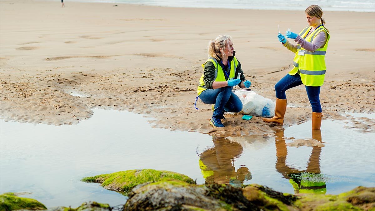 Two people wearing high-visibility vests take water samples from a shallow pool of water.