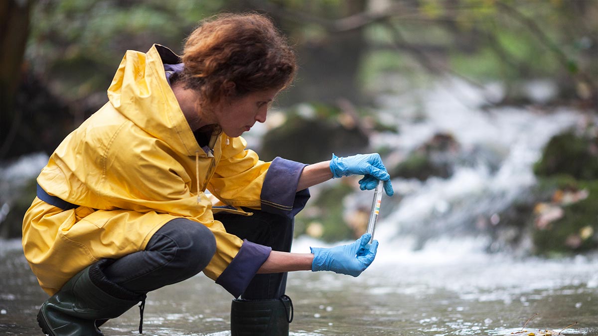 A person crouching in a river and testing the water with a small tube and a slip of paper with differed colored tabs.