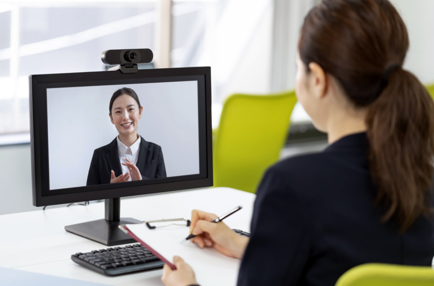 A woman sits with her back to the camera at a desk with a pen and clipboard in her hands, taking notes as she interacts with another woman on her computer screen