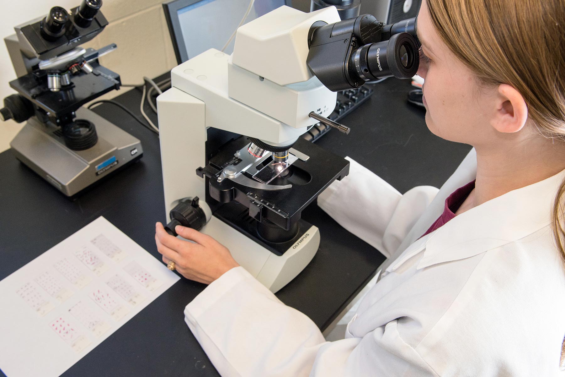 A female student wearing a white lab coat looks into the eyepiece of a microscope within a biology laboratory at Texas A&M University