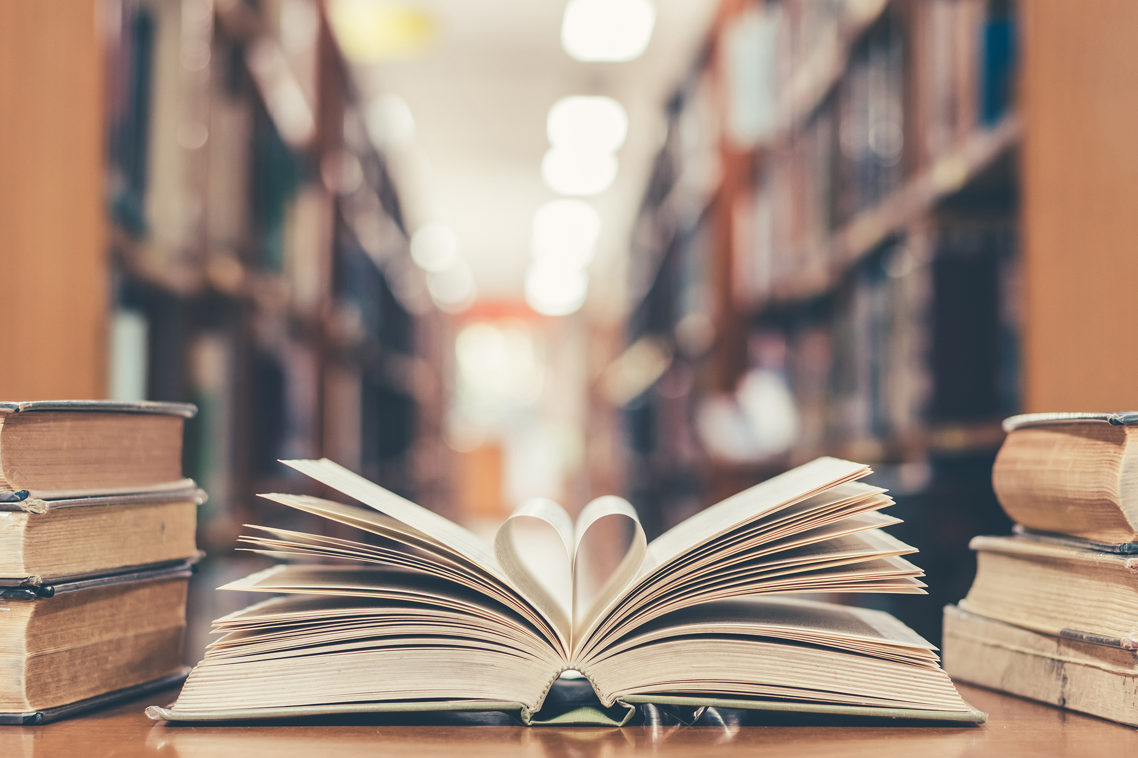 Book with open pages folded in a heart shape and stacked piles of textbooks on a reading desk in a library with stacks in the background