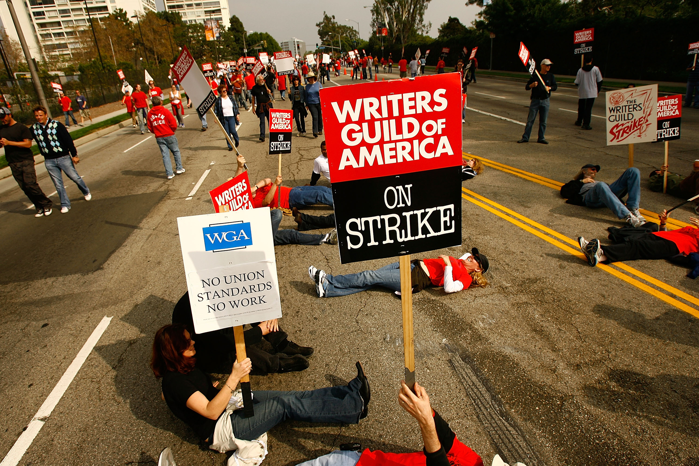 Writers Guild of America members and supporters, who are pictured standing, walking and lying down on a city street while holding protest signs in solidarity with the current writers' strike