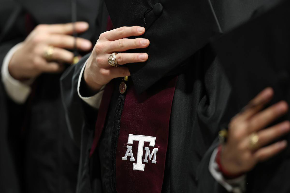 Close-up of Texas A&amp;M University graduates in regalia, holding their mortar boards with their Aggie Rings and maroon stoles featuring the white aTm logo visible