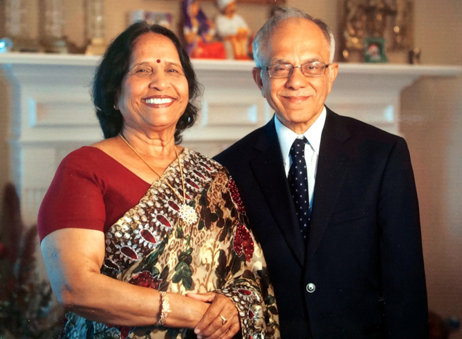 Two individuals smiling for the camera in a home setting, with a photo of Mahatma Gandhi in the background. One person is wearing a sari and the other is dressed in a suit.