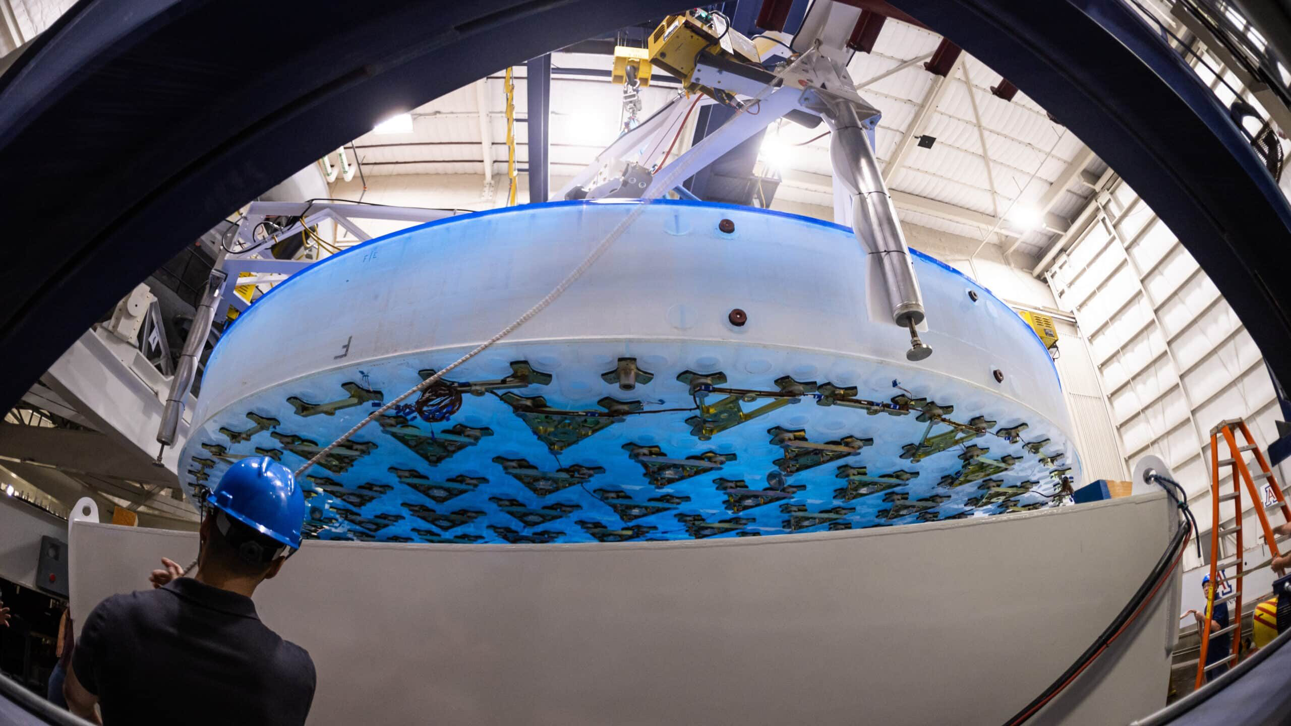 Completed 8.4-meter-diameter primary mirror for the Giant Magellan Telescope, hanging above the support system prototype with a technician in the foreground at the University of Arizona’s Richard F. Caris Mirror Lab