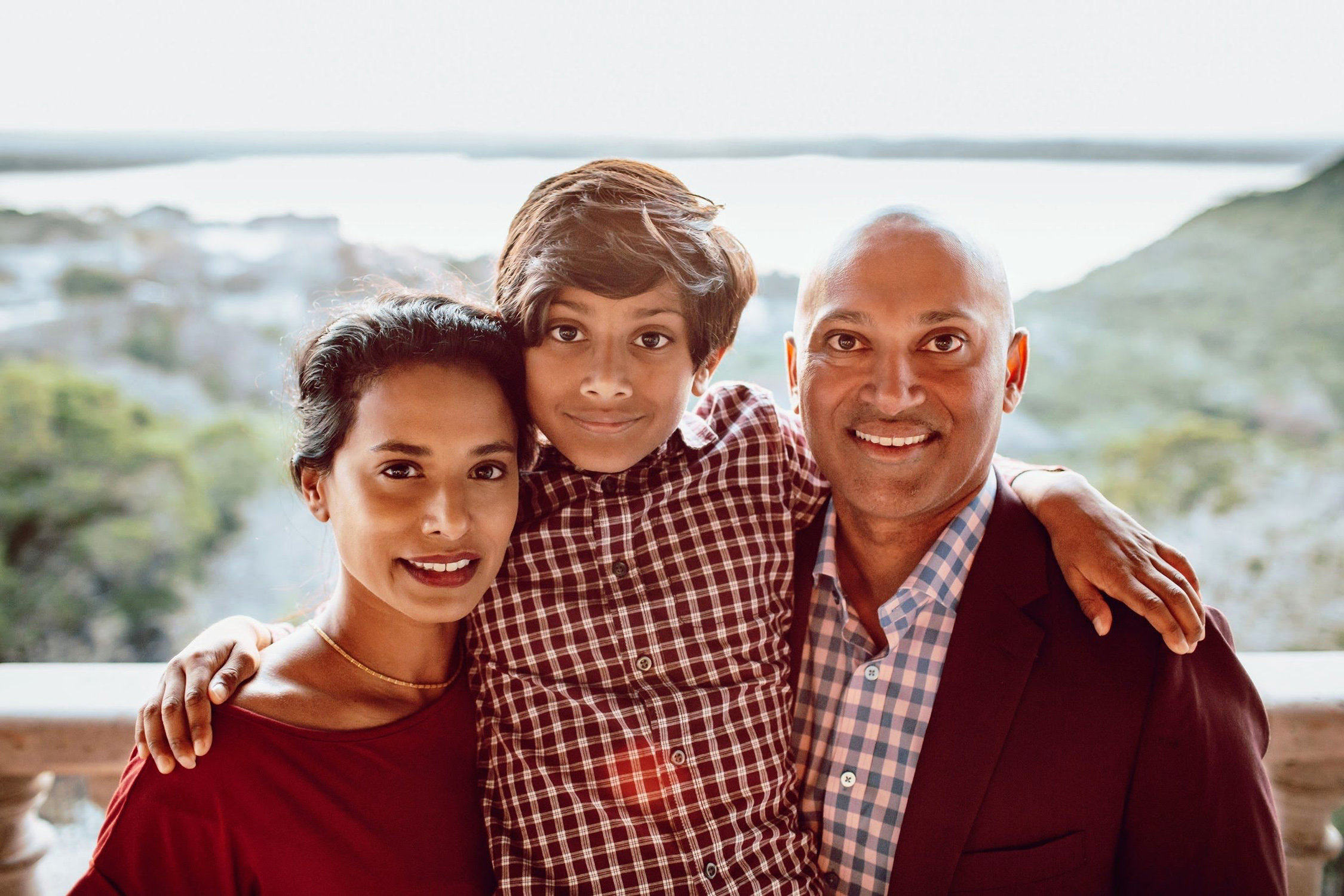 A family of three, featuring two adults and one child, smiling towards the camera with a scenic blurred background of hills and sky.