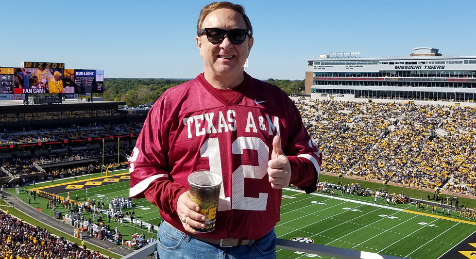 Person in a Texas A&amp;M jersey giving a thumbs-up at a football game between Missouri Tigers and Texas A&amp;M, with a stadium filled with spectators in the background.