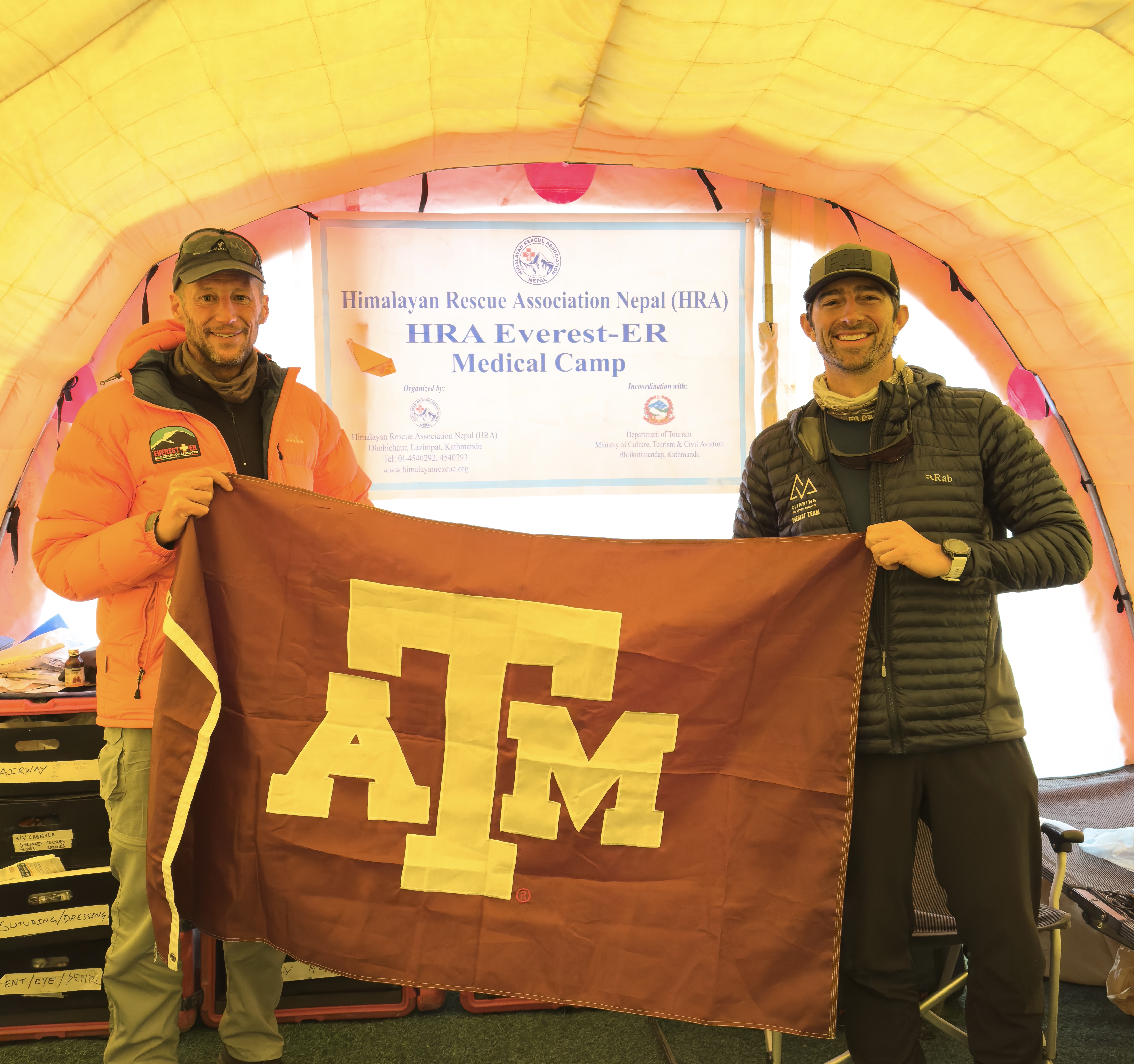 Two individuals holding a Texas A&M flag initials inside a tent at the Himalayan Rescue Association Nepal (HRA) Everest-ER Medical Camp.