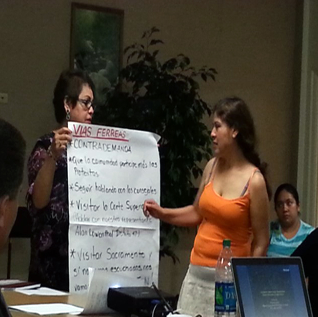 Two Latina women interacting during a training session -- the older one holding a poster-sized sheet of white paper outlining various tips in Spanish while the younger woman reads the information