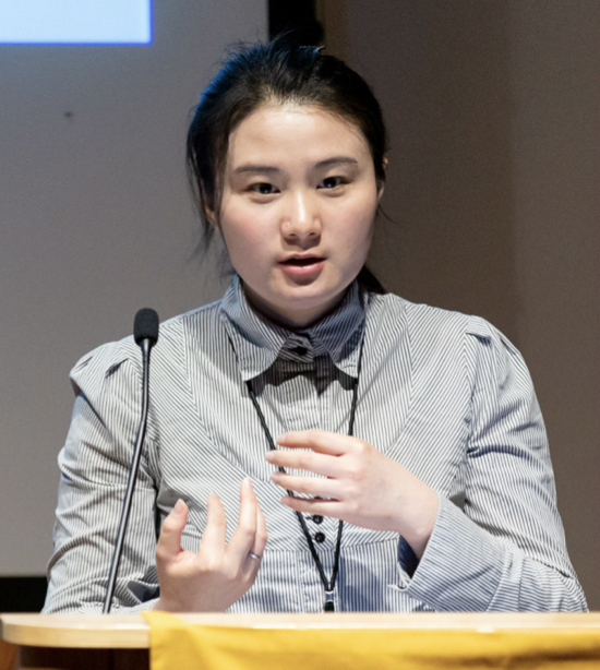 Dr. Zhang, wearing a striped blazer, is giving a presentation at a podium.