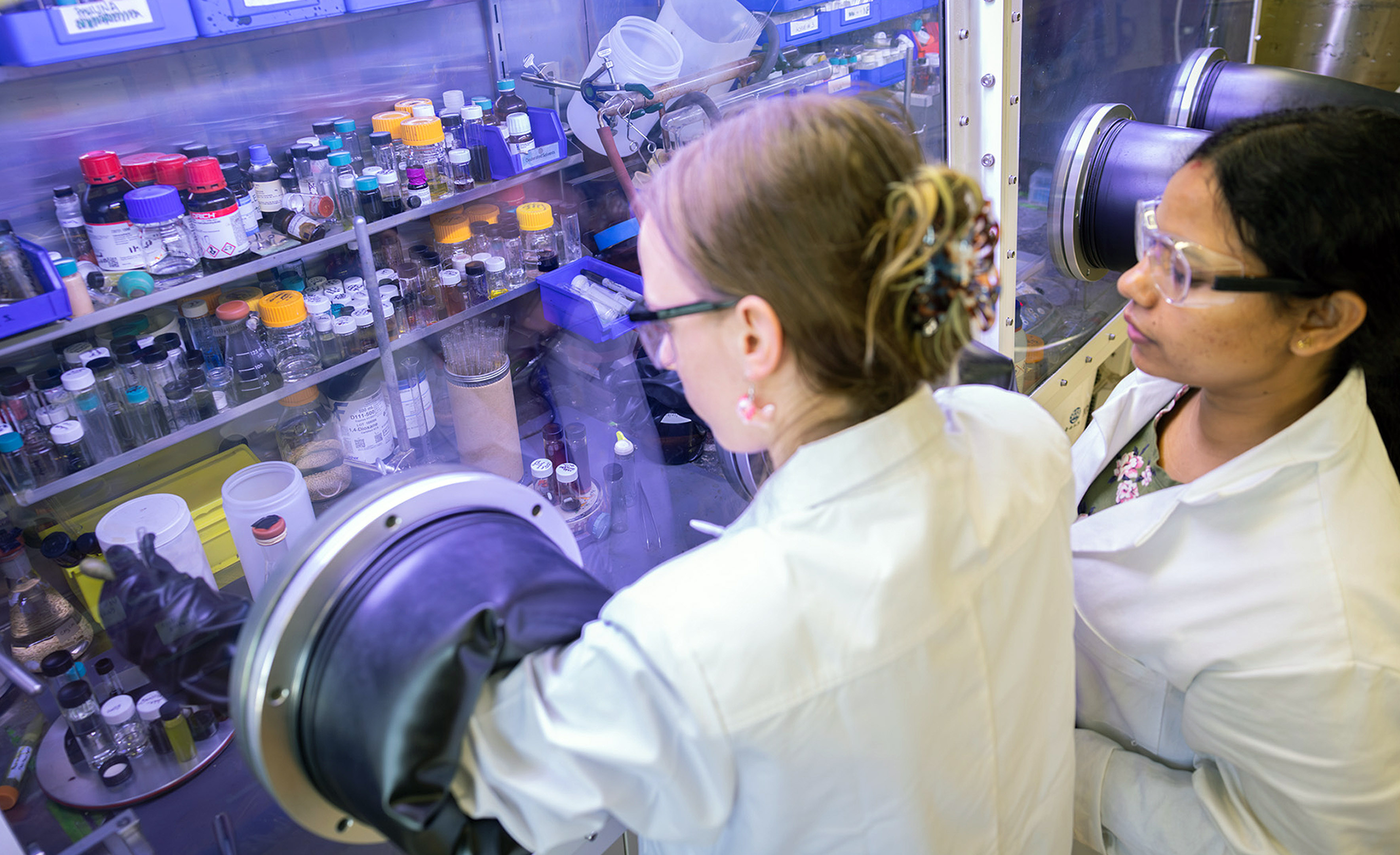 Two scientists working in a laboratory, one viewing a sample in a glove box and the other observing. The lab setting includes various scientific equipment and containers.