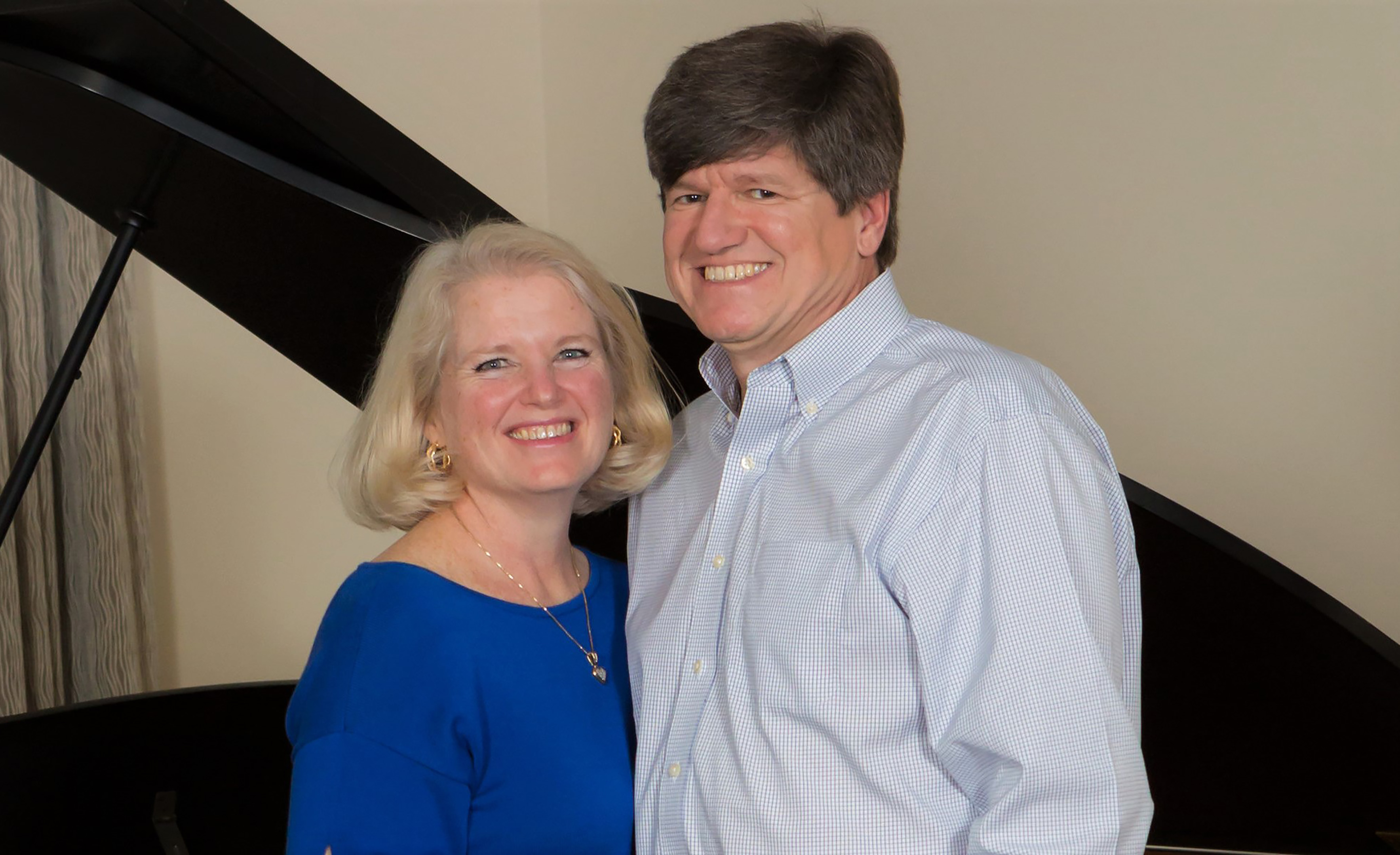 A man in a collared shirt smiling for a photo next to a blonde woman in a blue blouse. They're very happy and looking at the camera.