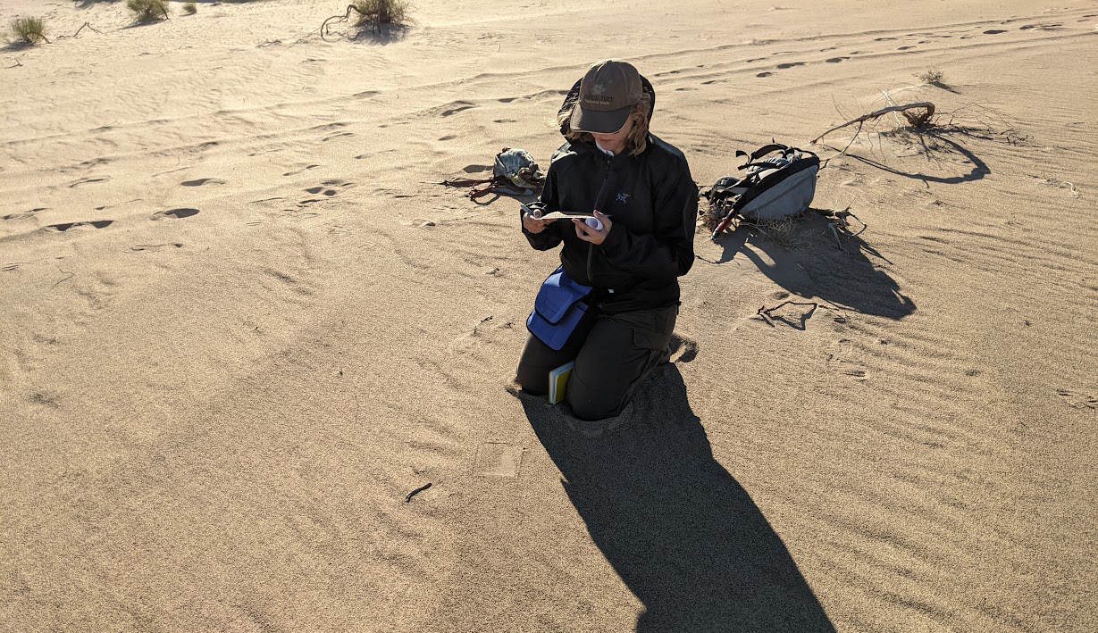 Lauren Berger is crouched on a sandy surface operating a drone remote control with a backpack beside them.