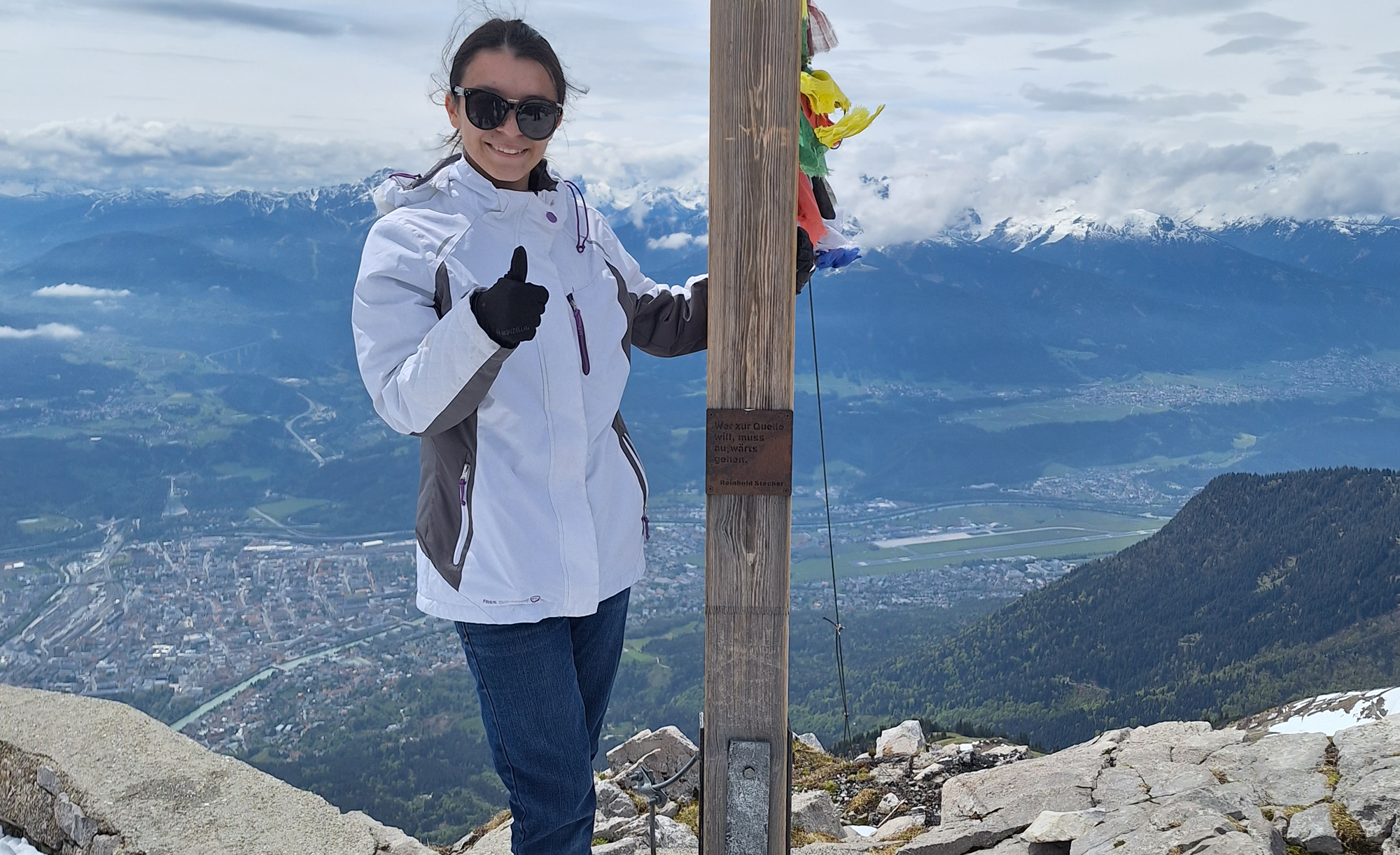 Juliette Rocha standing next to a wooden post with prayer flags, giving a thumbs-up, with a panoramic view of a mountain range and a city below in the background.