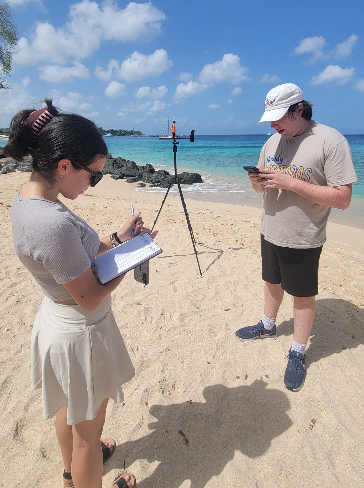 Juliette Rocha (left) and Brian Neuville (right) are standing on a sandy beach, each using a smartphone. Rocha is also holding a notebook. A tripod with a device is set up beside them, with the ocean and a clear blue sky in the background.