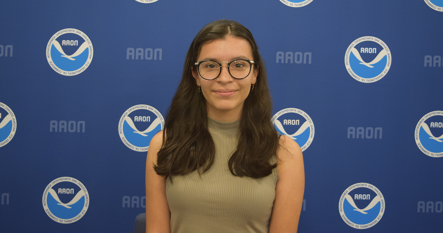 Juliette Rocha with glasses, wearing a green shirt, standing in front of a blue backdrop featuring the AAON logo.