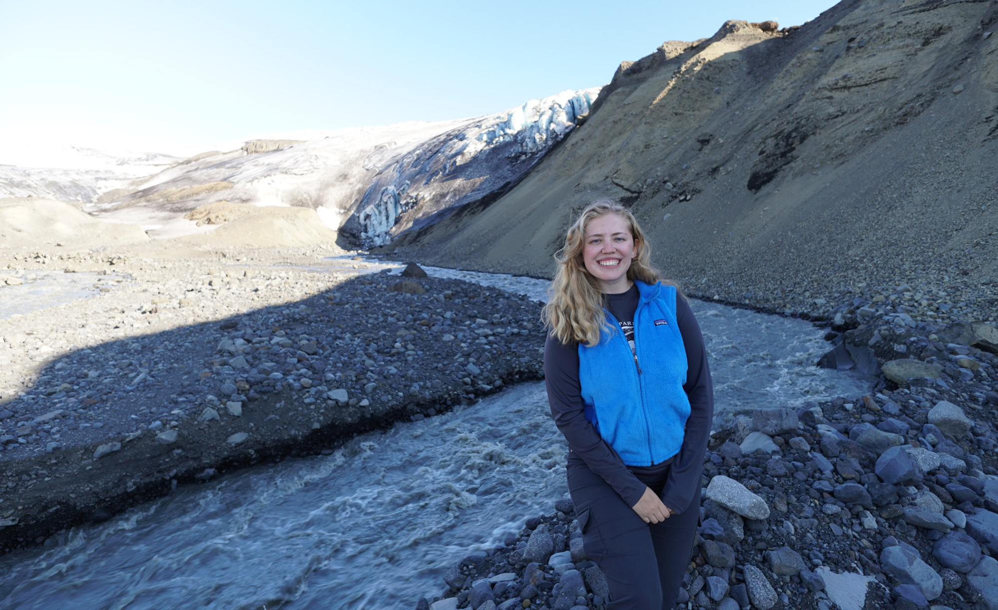 Lauren Berger is standing in a blue jacket smiling in front of a scenic view with a flowing river and a glacier in the background.