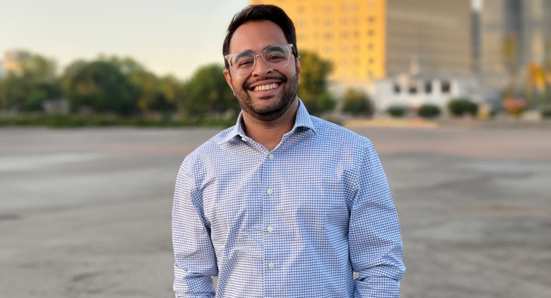 Sahir Gagan smiling with glasses, wearing a blue checkered shirt, standing outdoors with city buildings in the background.