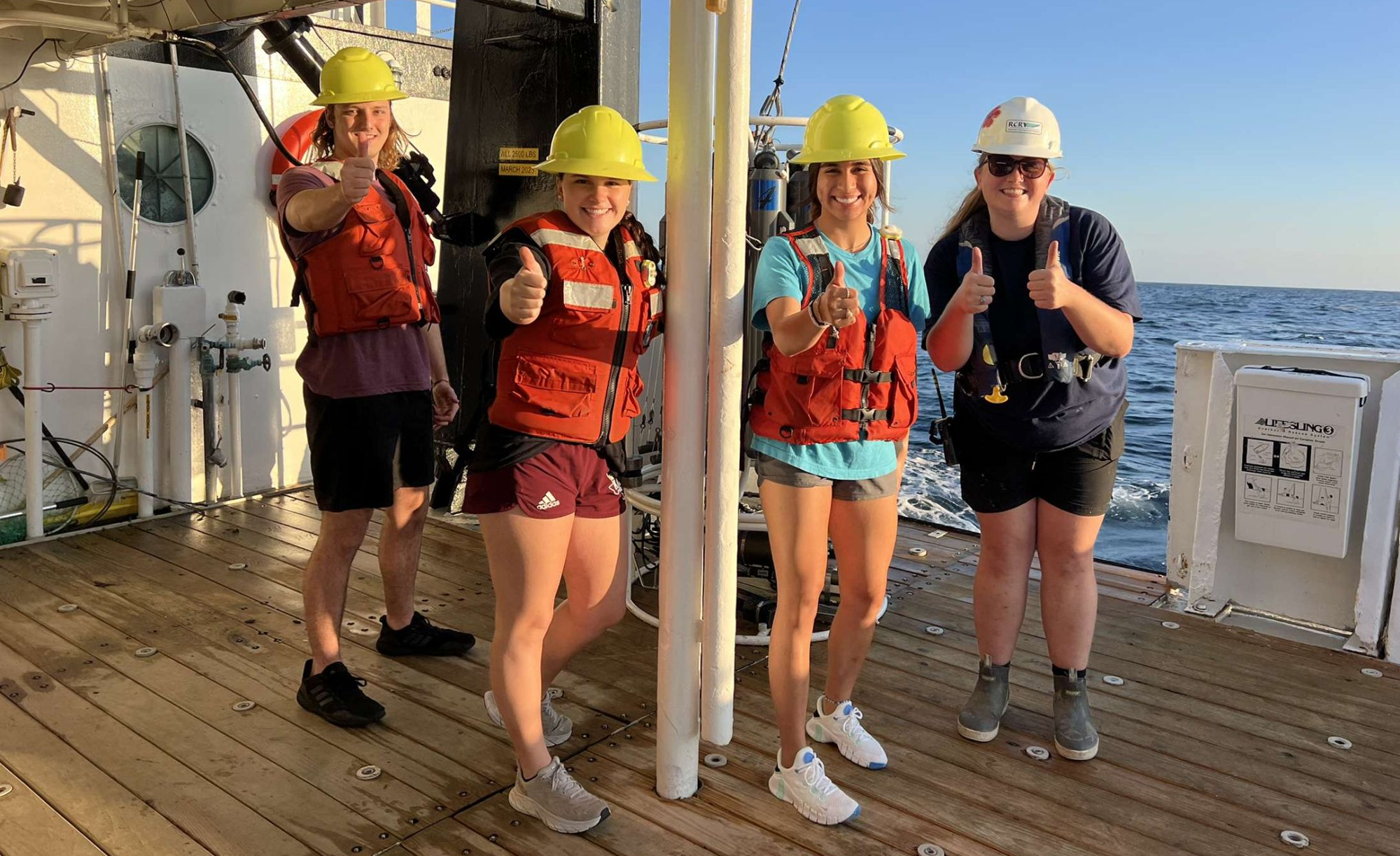 Four individuals wearing safety gear including helmets and life jackets, giving thumbs up on the deck of a ship at sea.