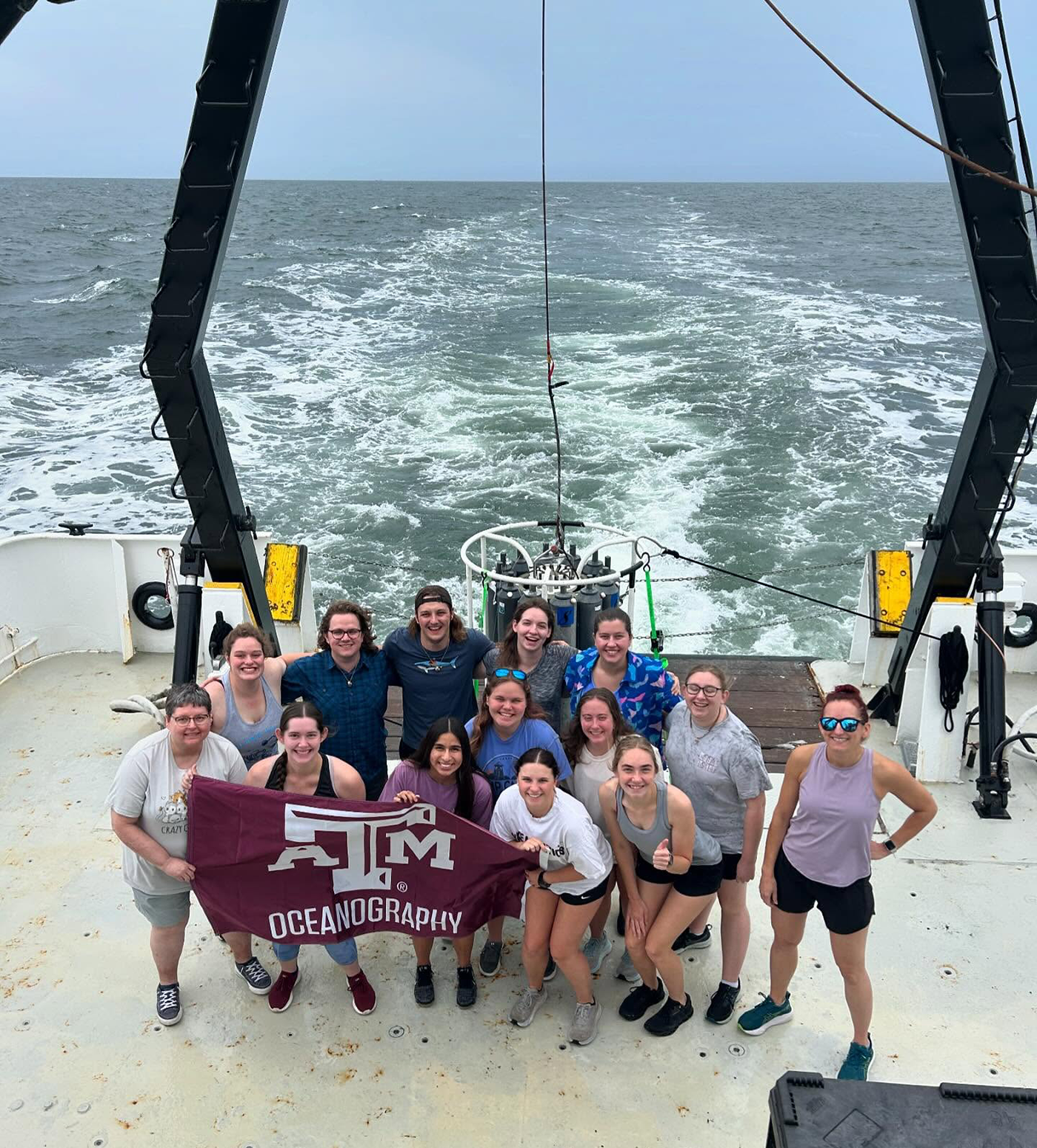 Group of students and faculty holding a Texas A&amp;M Oceanography banner on the deck of a boat with the ocean in the background.