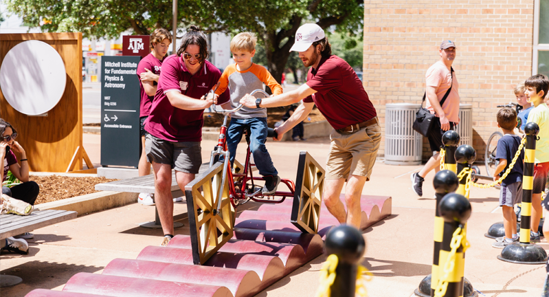 Two adults assist a child riding a bicycle with squae wheels on an elevated, wavy track at the Texas A&M 2023 Physics Festival, with other spectators in the background.