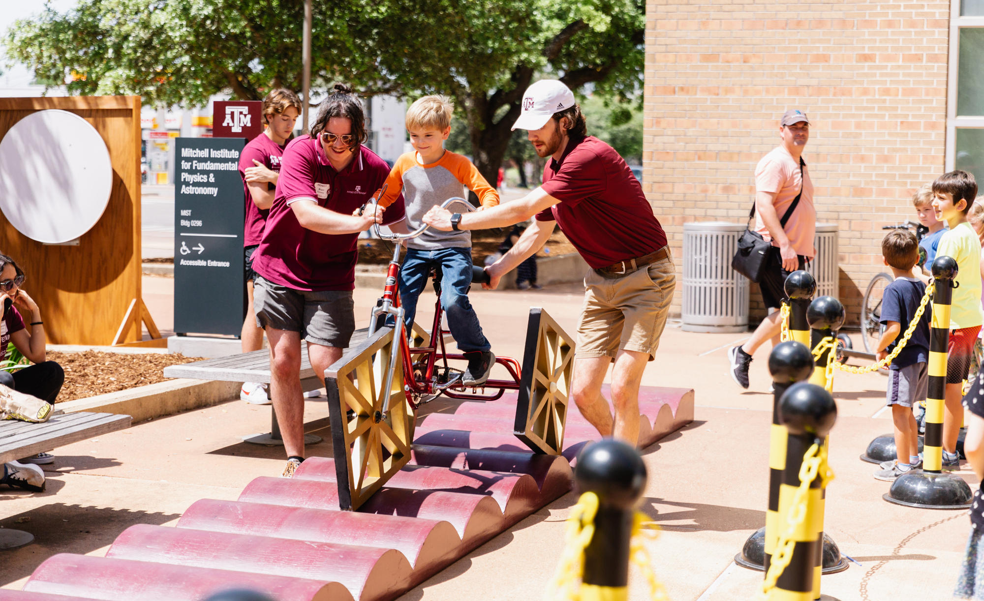 Two adults assist a child riding a bicycle with squae wheels on an elevated, wavy track at the Texas A&amp;M 2023 Physics Festival, with other spectators in the background.