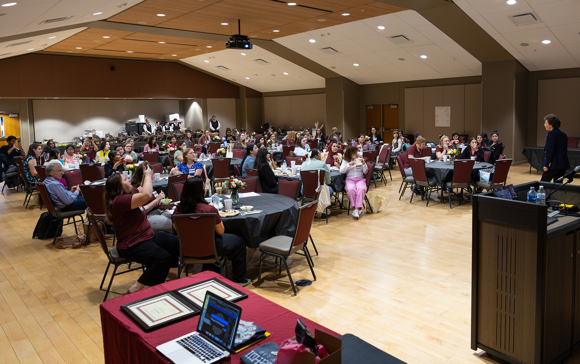 Attendees seated at round tables listen to a speaker at the front of a spacious conference hall.