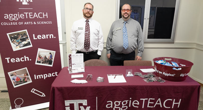 Kenric Davies and Colby Richardson standing behind a promotional table for the AggieTEACH program at Texas A&M University, flanked by banners displaying the university's logo and the College of Arts & Sciences. The table features brochures and promotional items.