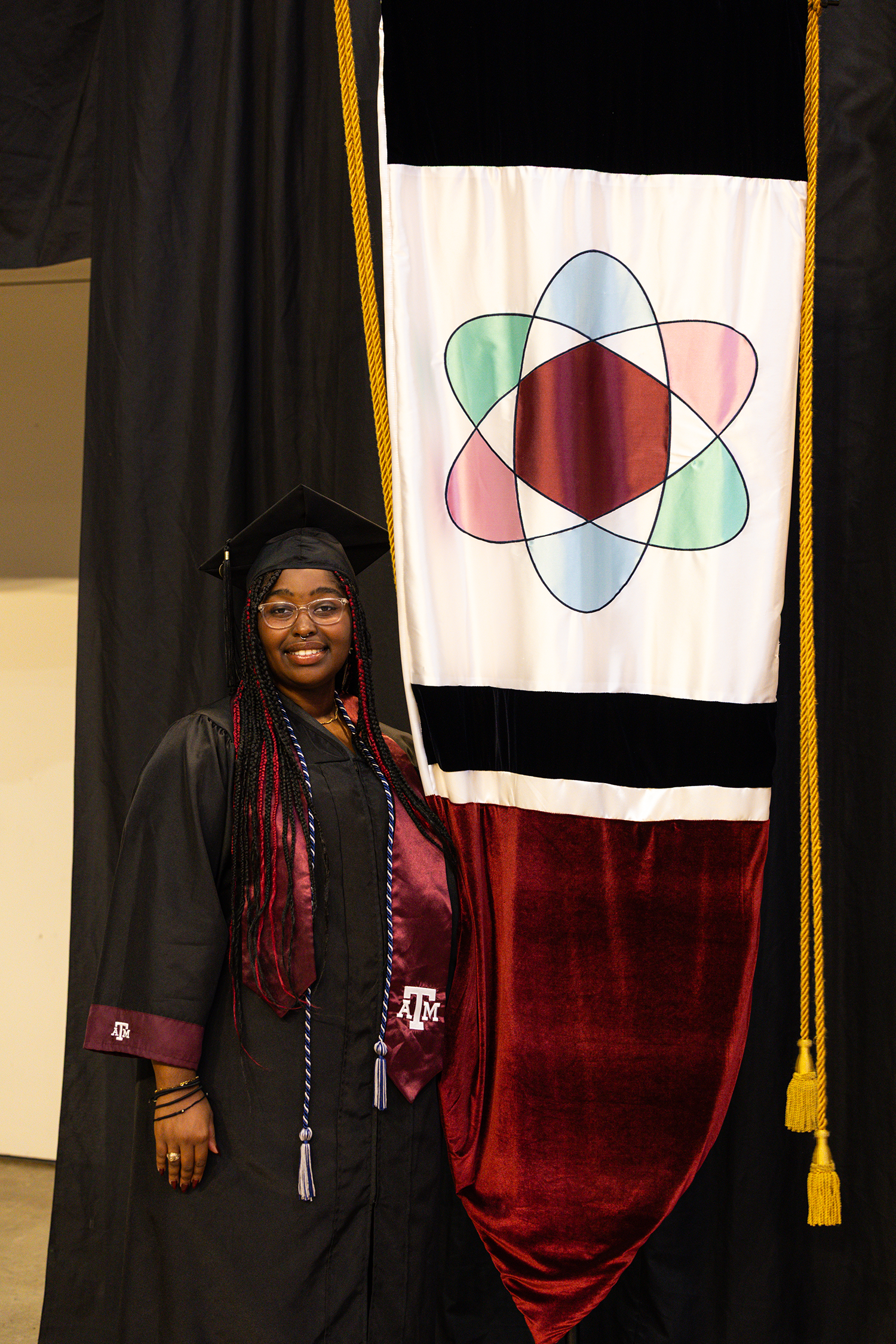 A graduate in cap and gown stands beside a Texas A&amp;M University banner, smiling proudly.