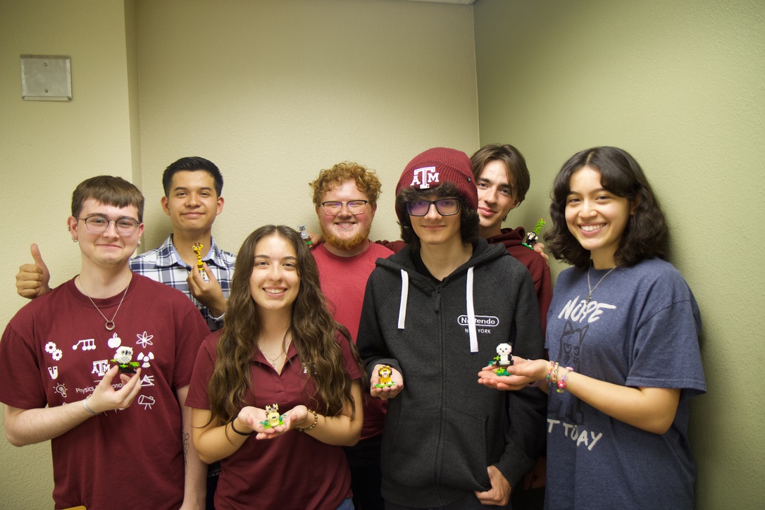 Group of seven individuals in a room, smiling and holding colorful puzzle cubes. Some are wearing shirts with logos including Texas A&amp;M and Google.