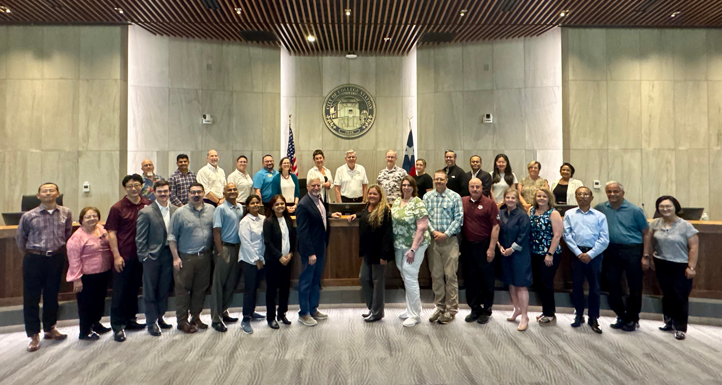 Group of people standing in a city council chamber in front of the official seal and flags.