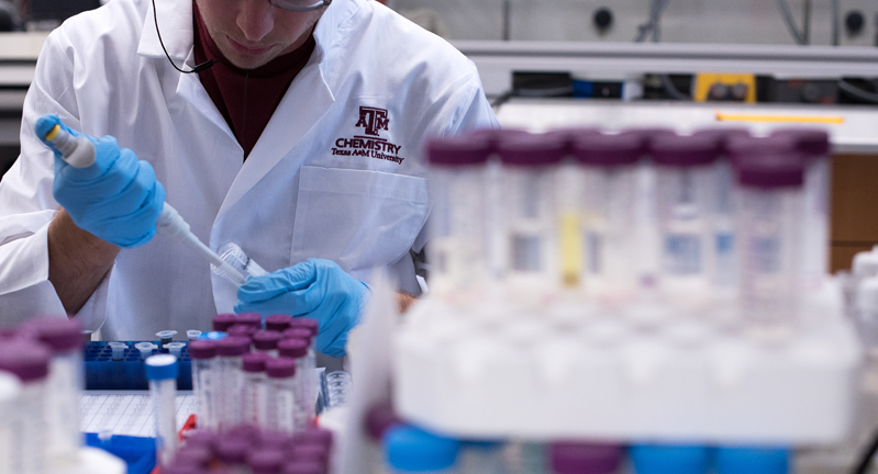 A Texas A&amp;M University lab technician in a white lab coat labeled "Chemistry" is pipetting a substance into a test tube, surrounded by racks of samples.