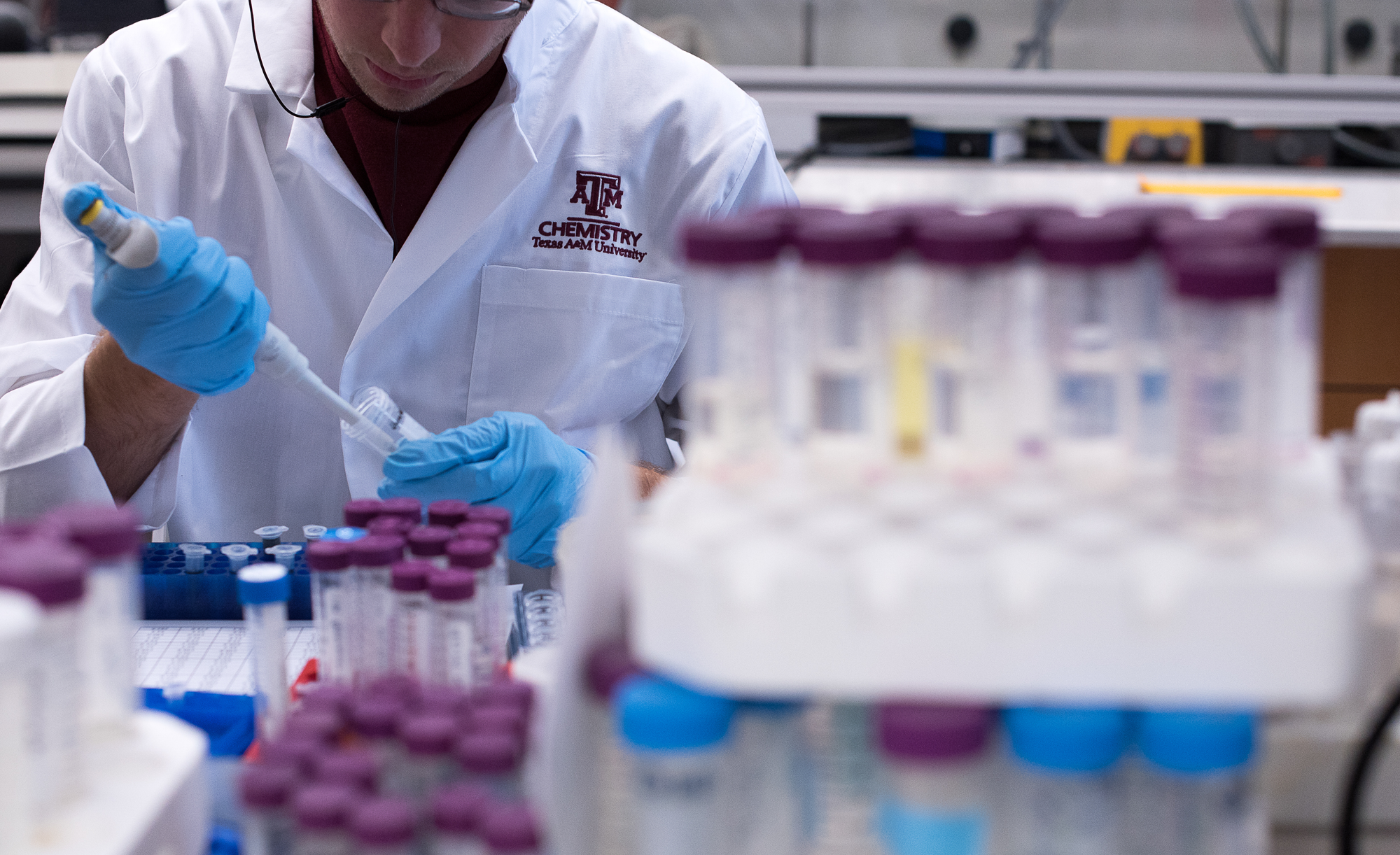 A Texas A&M University lab technician in a white lab coat labeled "Chemistry" is pipetting a substance into a test tube, surrounded by racks of samples.