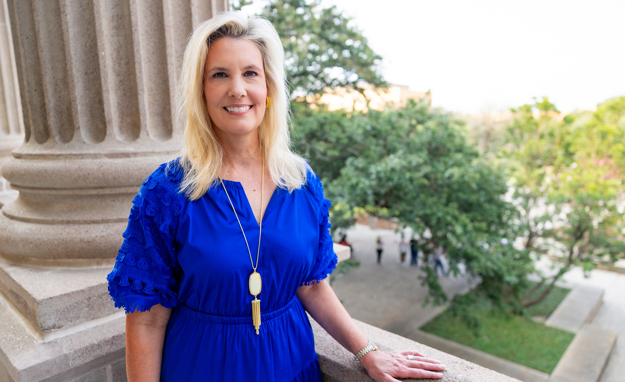 Image of a blonde woman in a blue dress on a balcony overlooking the Century Tree, in front of a concrete pillar with her hand on the railing.