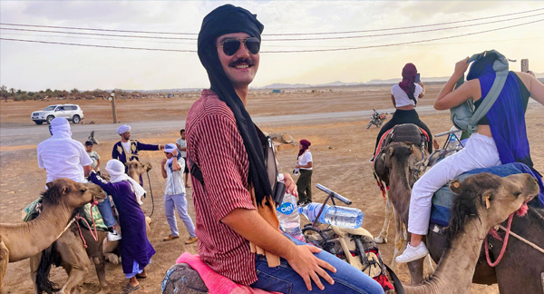 Person smiling while sitting on a camel, with other riders and camels in the background under a clear sky.