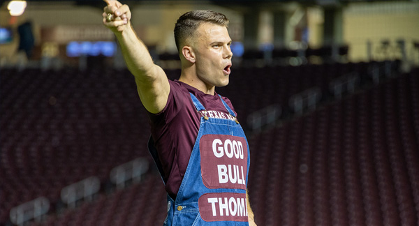 A person wearing a maroon shirt and overalls that say "Good Bull" and "Thomas" leads Midnight Yell practice.
