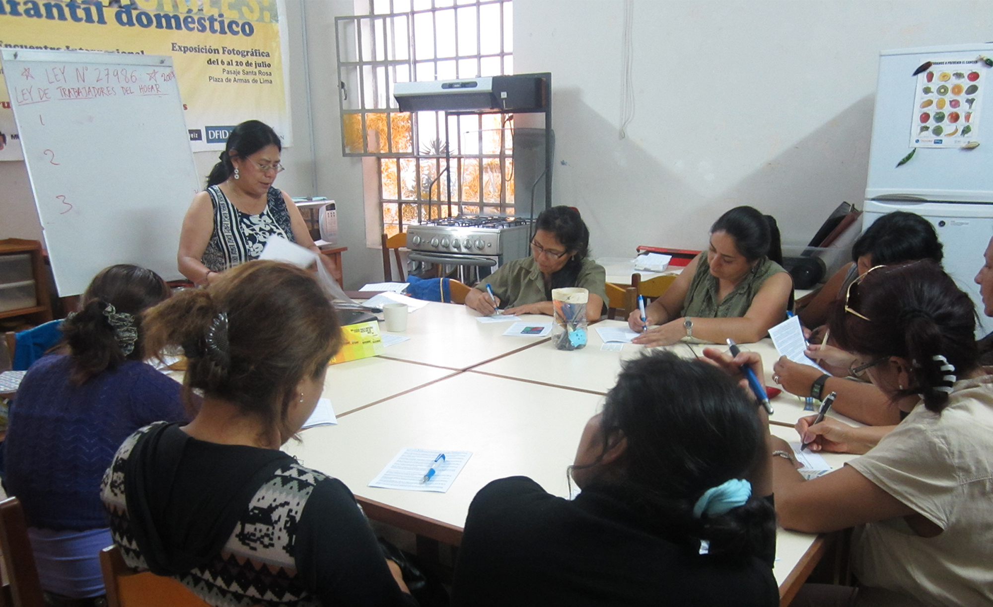 A group of people seated around a rectangular table in a classroom setting, writing on papers while one person stands at the front holding documents near a whiteboard.