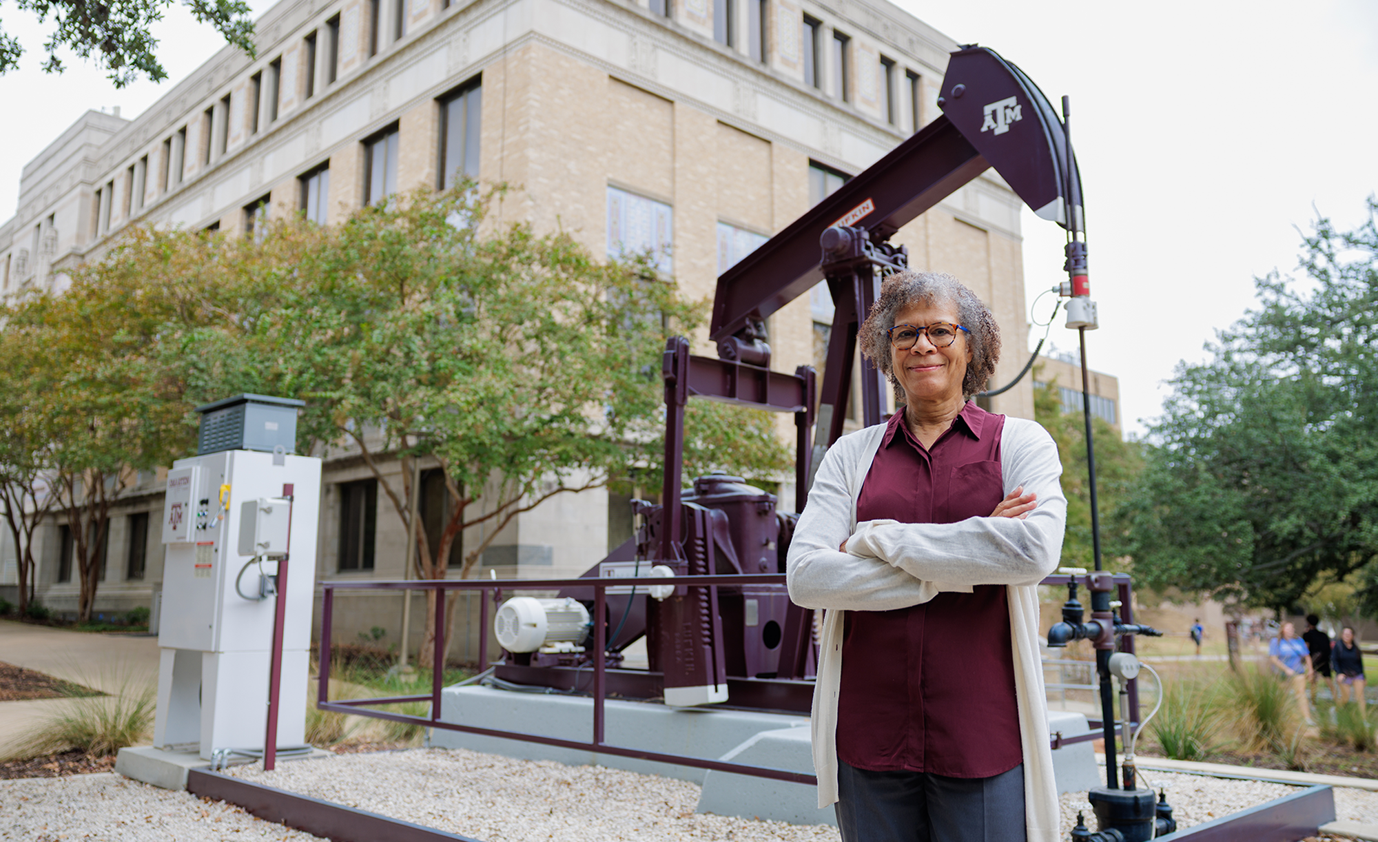 A person stands with arms crossed in front of a large oil pump jack bearing the Texas A&amp;M University logo, set on a university campus with a beige brick building and surrounding greenery in the background