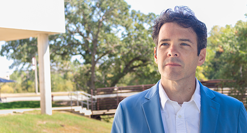 Jonathan Meer, economics department head at Texas A&amp;M University, wearing a light blue blazer and white shirt, standing outdoors on campus with trees, a pedestrian bridge, and part of a white building in the background.