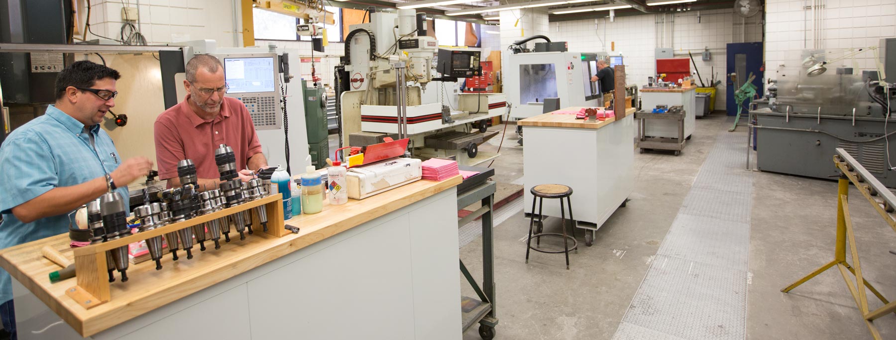 Garrick Garza and John Lopes working at table in the machine shop. The table has machined parts and chemicals in bottles. Machine shop equipment is in the background. 